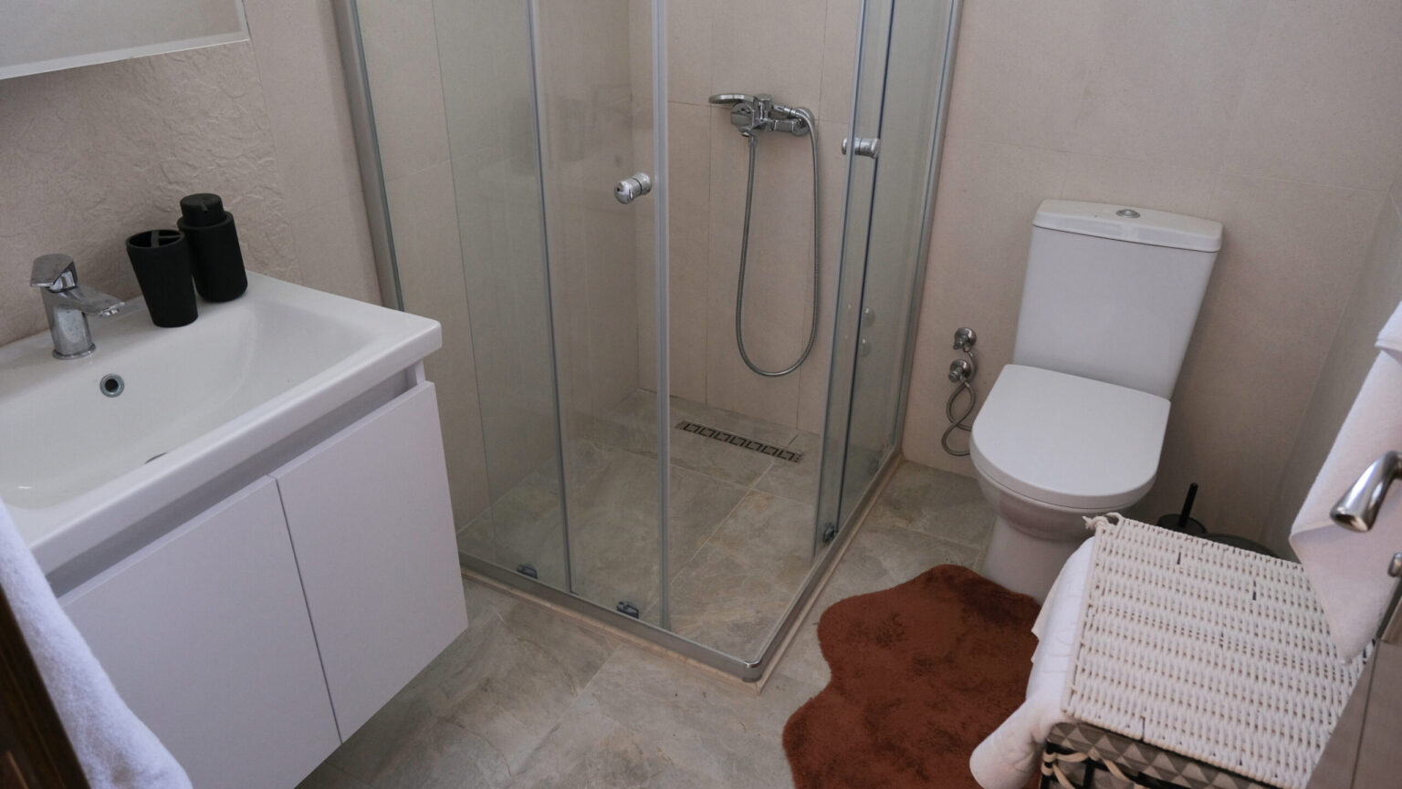 A modern bathroom with a glass shower enclosure, white sink cabinet, toilet, brown rug, and a wicker laundry basket.