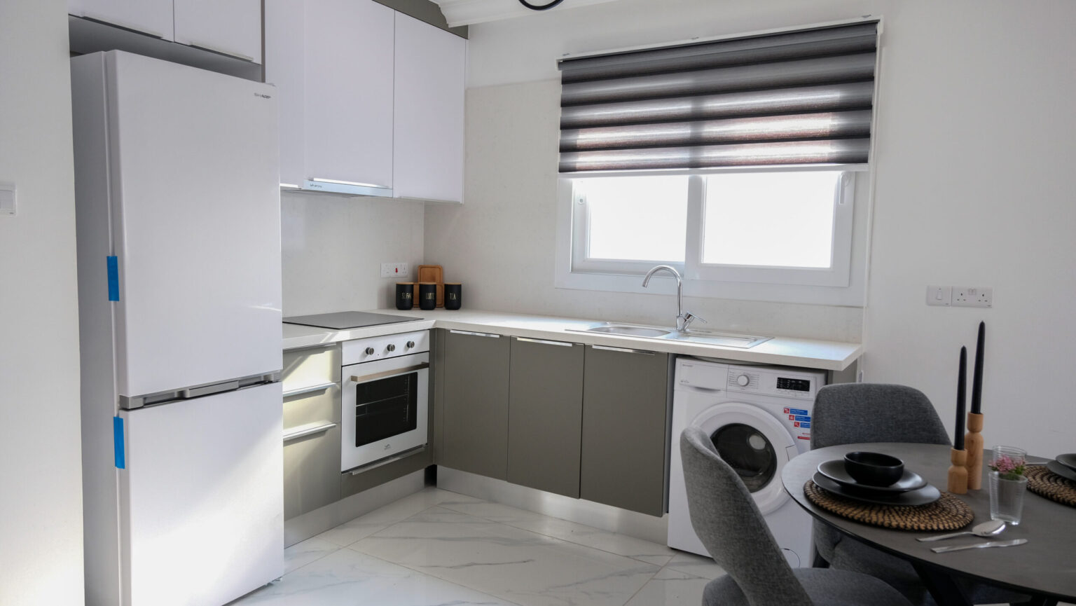 Modern kitchen with a white fridge, oven, and washing machine, L-shaped counters, gray chairs around a dining table, and a window with a striped blind.