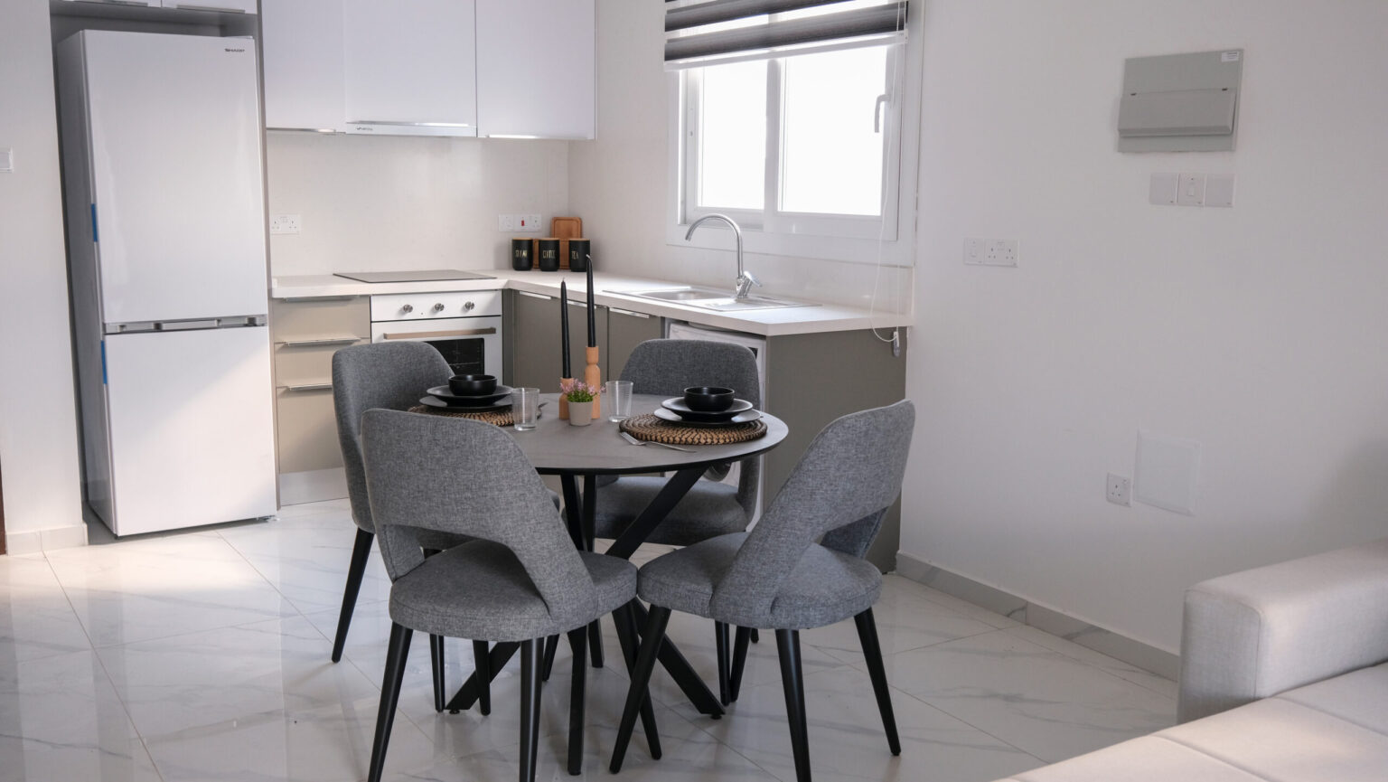 A modern kitchen and dining area with a round table set for four, gray chairs, white cabinets, a refrigerator, and a window above the sink.
