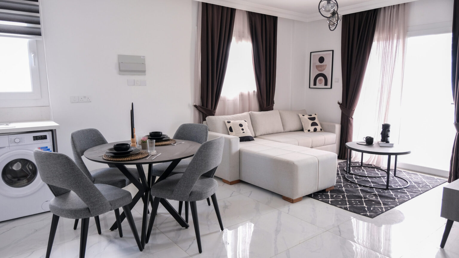 Modern living and dining area with gray chairs, a round table set for four, a light-colored sectional sofa, and dark curtains; washing machine visible in the corner.