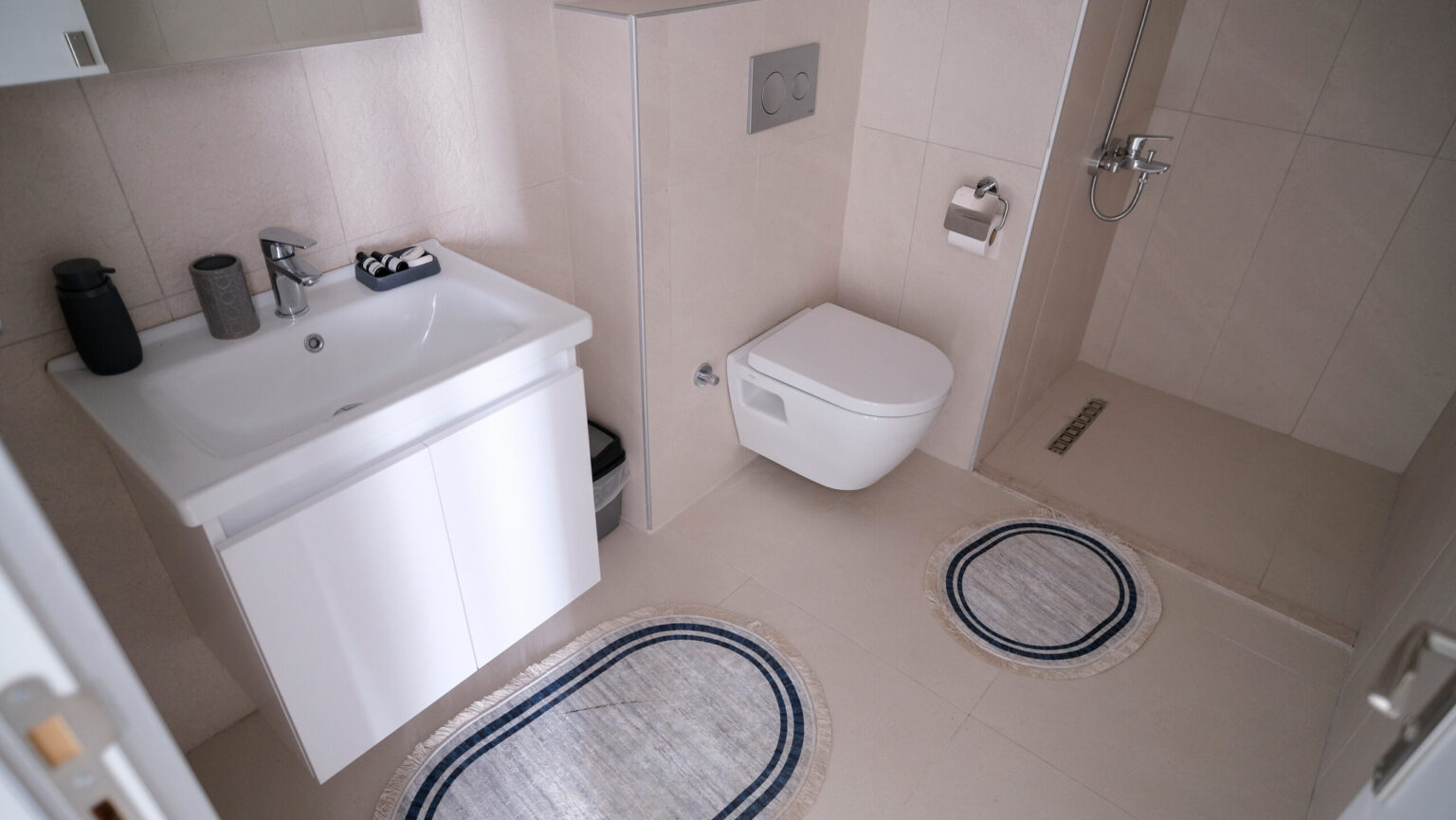 Modern bathroom with a wall-mounted toilet, white sink cabinet, walk-in shower, and two oval rugs on a light tiled floor.