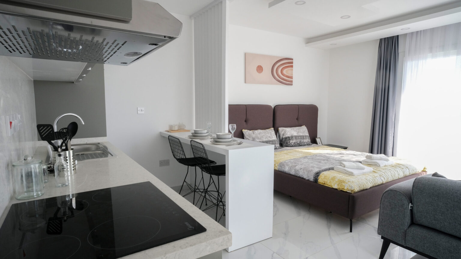 Modern studio apartment with a bed, small dining area with two chairs, a kitchen with stove and sink, and large window with sheer curtains.