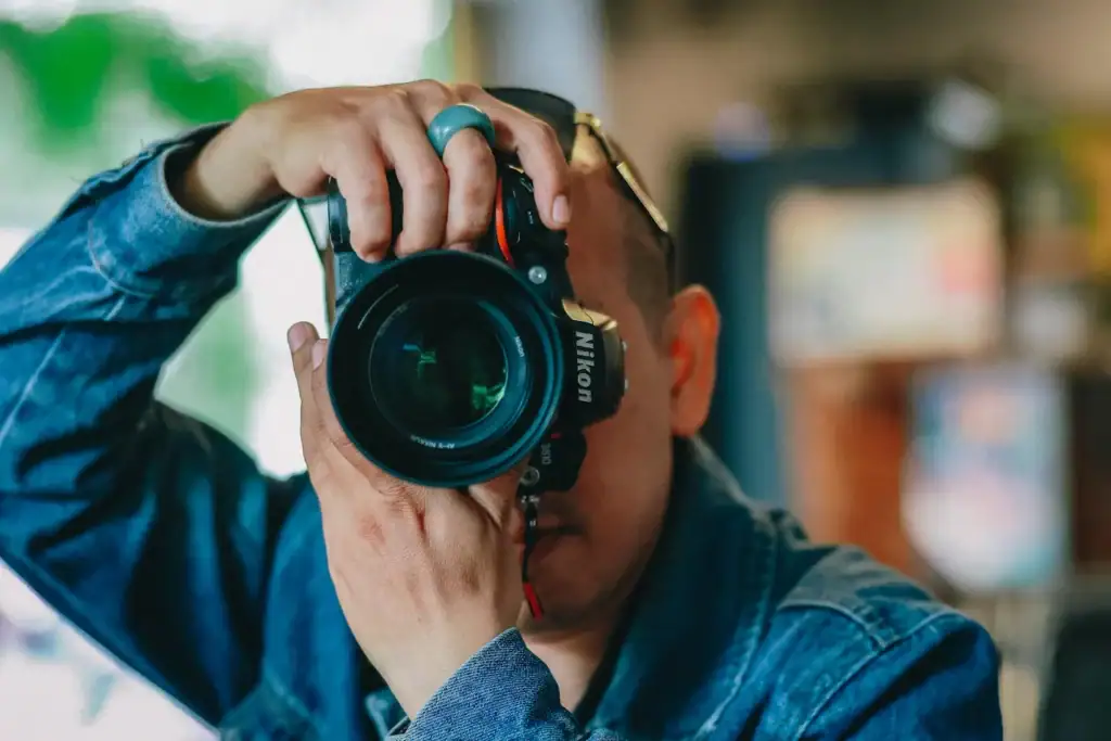 Person wearing a denim jacket holding a Nikon camera up to their face, capturing the vibrant atmosphere of a service de cuisine indoors.