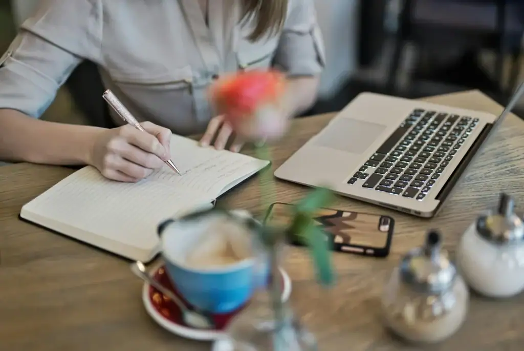 Person writing in a notebook beside a laptop, smartphone, and coffee cup on a wooden table with blurred flowers in the foreground—perfect for planning your next Service de cuisine experience.