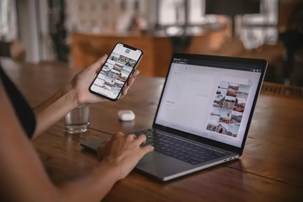 Person using a smartphone and laptop at a wooden table, managing photo content on both devices; a glass of water, wireless earbuds, and notes about a service de cuisine are nearby.