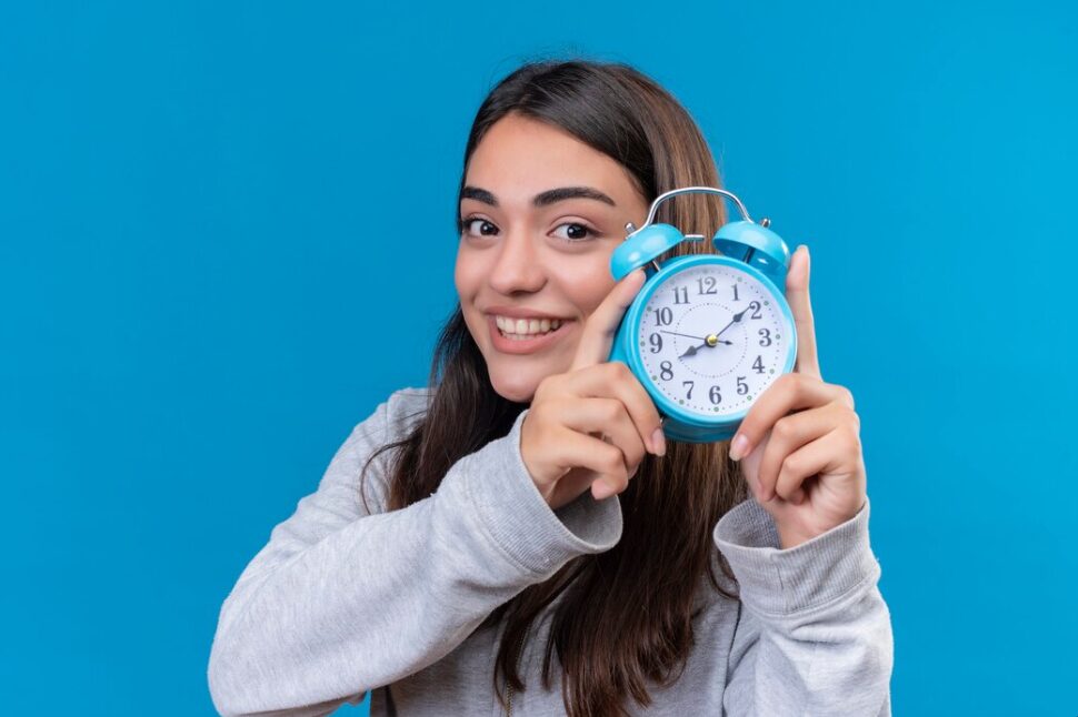 A young woman in a gray sweatshirt smiles while holding a blue analog alarm clock against a solid blue background.