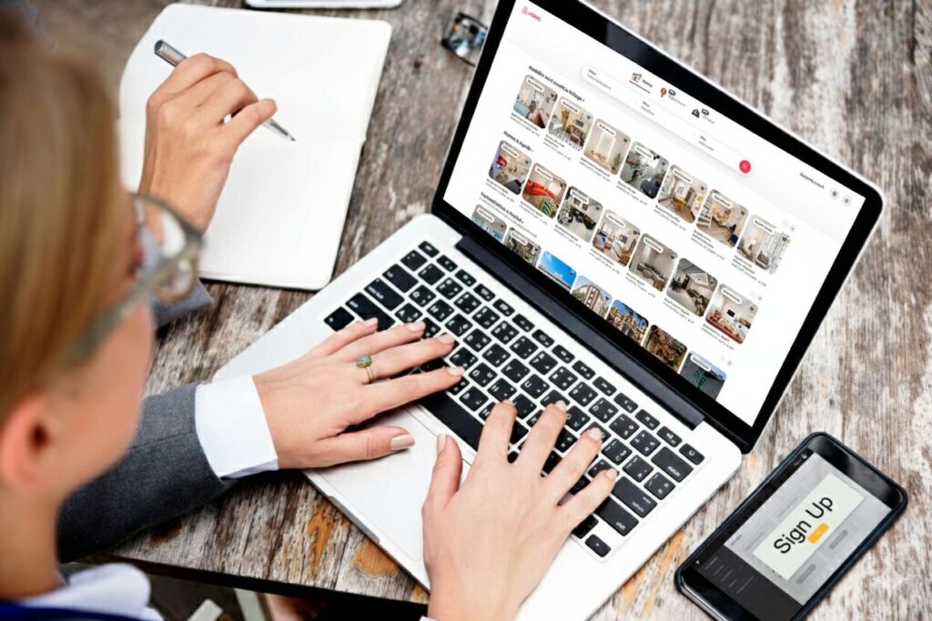 Person using a laptop to browse an image gallery website, with a notebook, pen, and a phone displaying a "Sign Up" screen on a wooden desk.