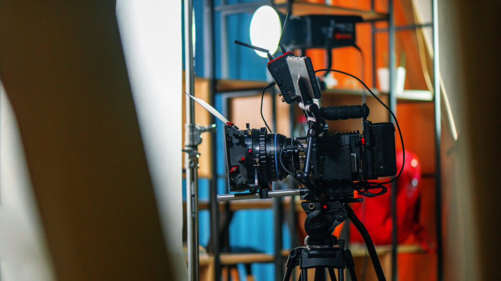 A professional video camera mounted on a tripod is set up in a brightly lit studio with shelves and various equipment in the background.